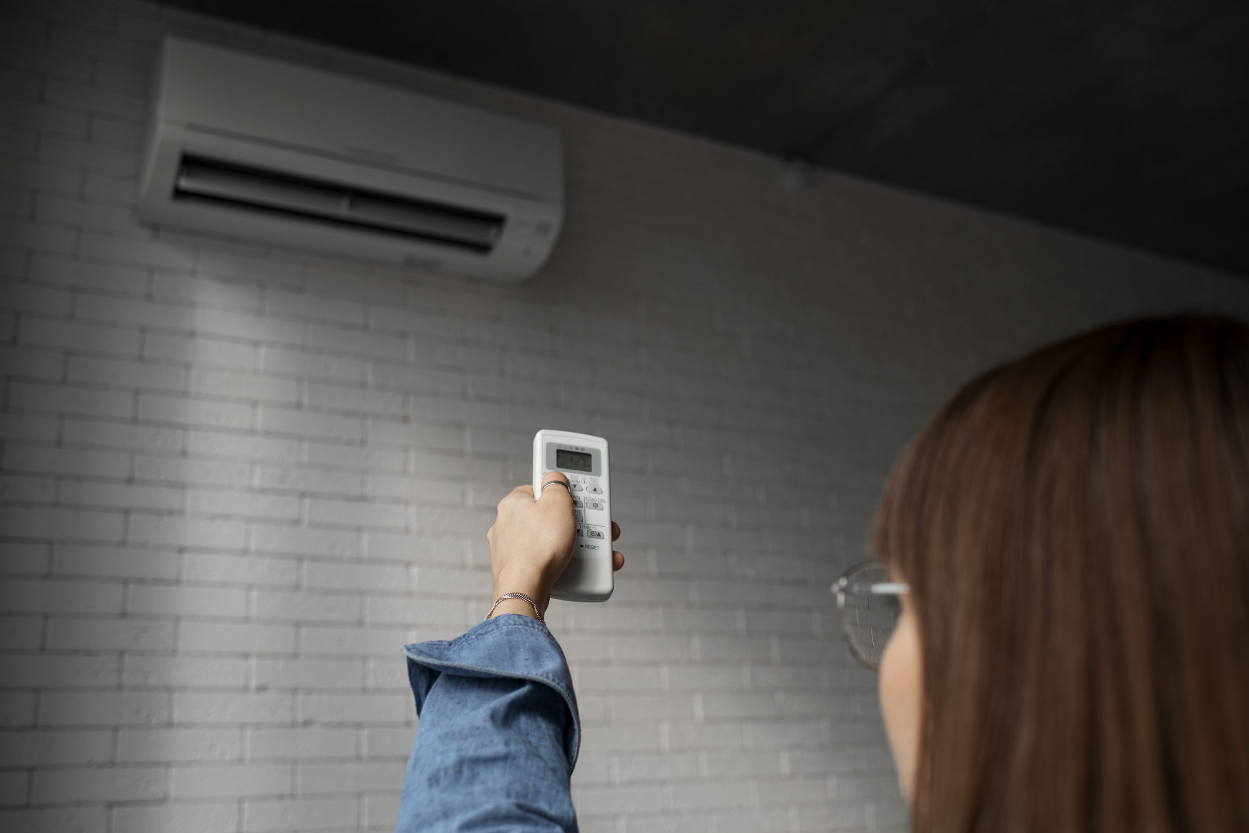 Mulher usando controle remoto para ajustar o ar-condicionado split na parede, com o aparelho em modo FAN ligado em ambiente interno iluminado.
