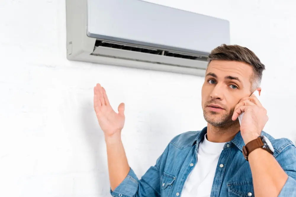 Homem segurando celular no ouvido enquanto aponta para ar condicionado com expressão tensa