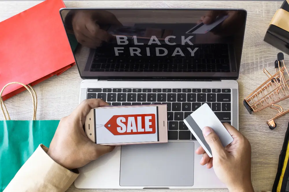 Visão de cima de pessoa segurando cartão com celular e notebook pós oferta ar-condicionado black friday 